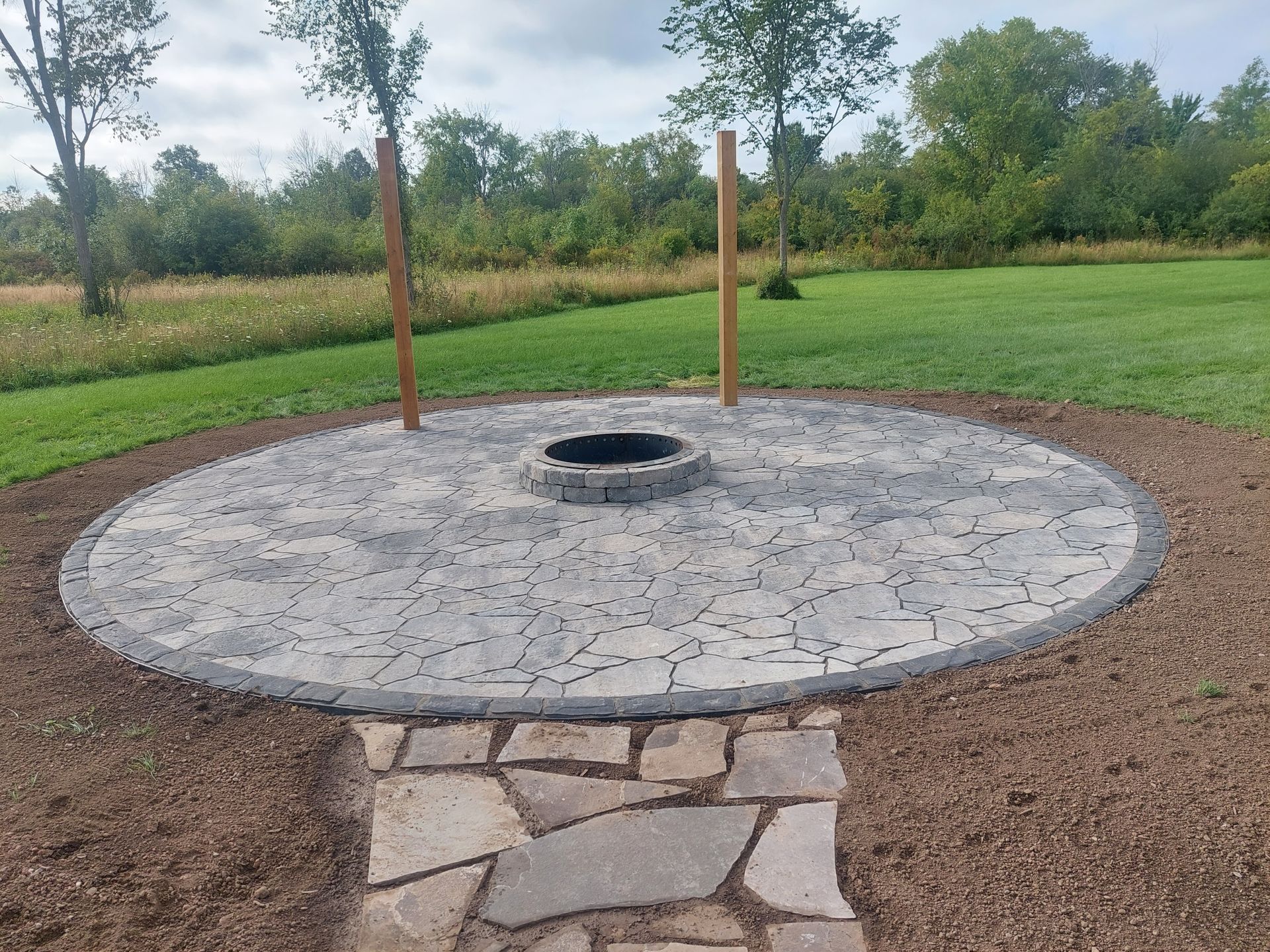 A circular stone patio with a central fire pit, two wooden posts, and a stone path in a grassy field.