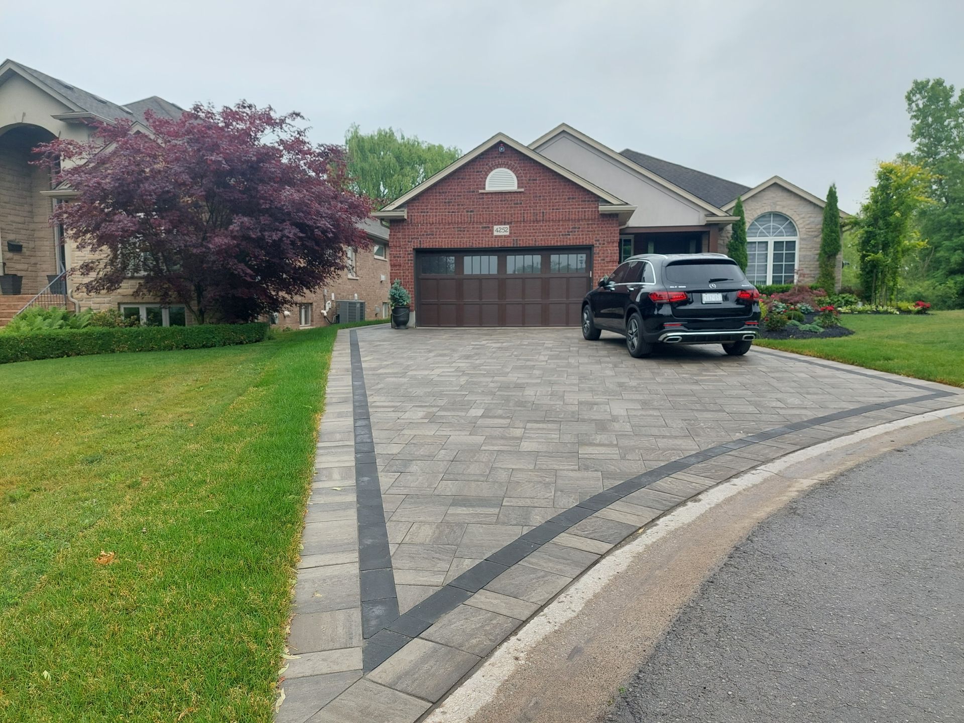 A brick house with a dark car parked in a paved driveway featuring a geometric border, next to a tree with red leaves.