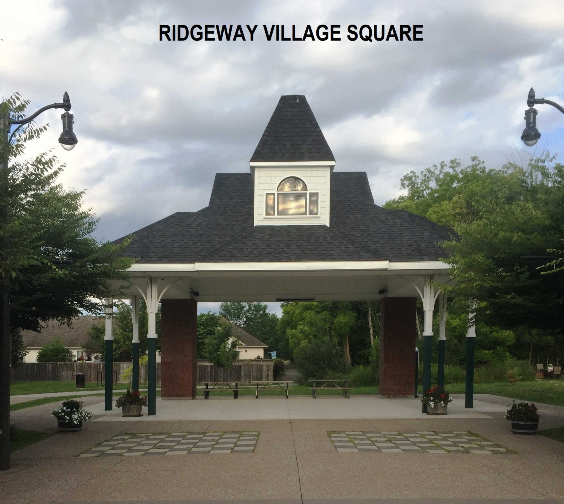 A pavilion at Ridgeway Village Square with a black roof, white trim, and brick columns under a cloudy sky.