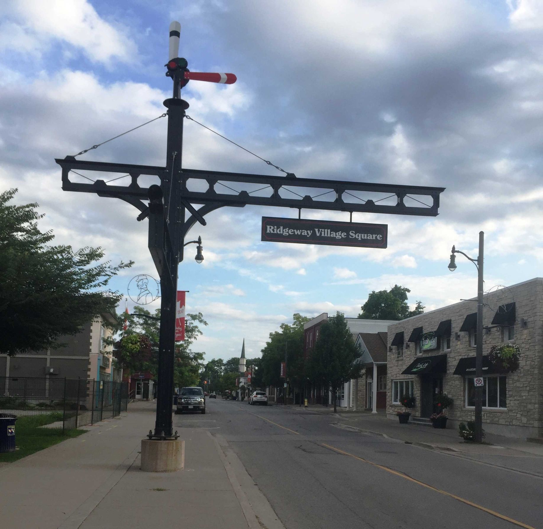 A sign for Ridgeway Village Square hangs from a historic railway-style signal post over a quiet street with brick buildings.