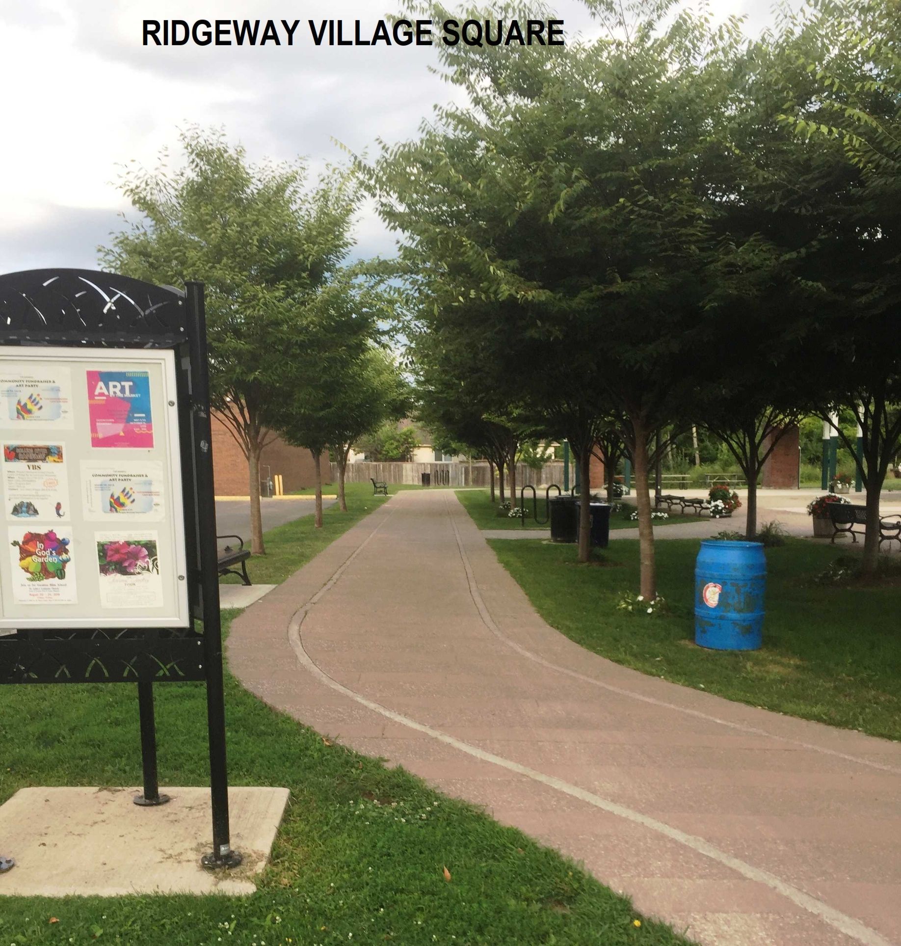 A paved walkway through Ridgeway Village Square, featuring an information kiosk on the left and trees along a grassy area.