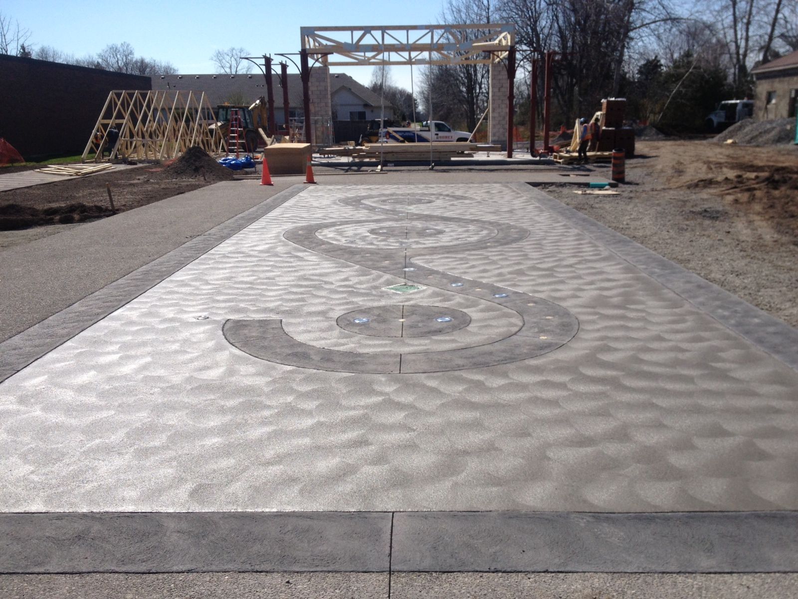 A stamped concrete driveway featuring a large, dark circular S-pattern inlay, located at an outdoor construction site.