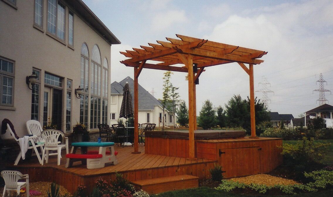 A wooden pergola stands over a hot tub on a raised deck next to a house, with white patio furniture nearby on the lawn.