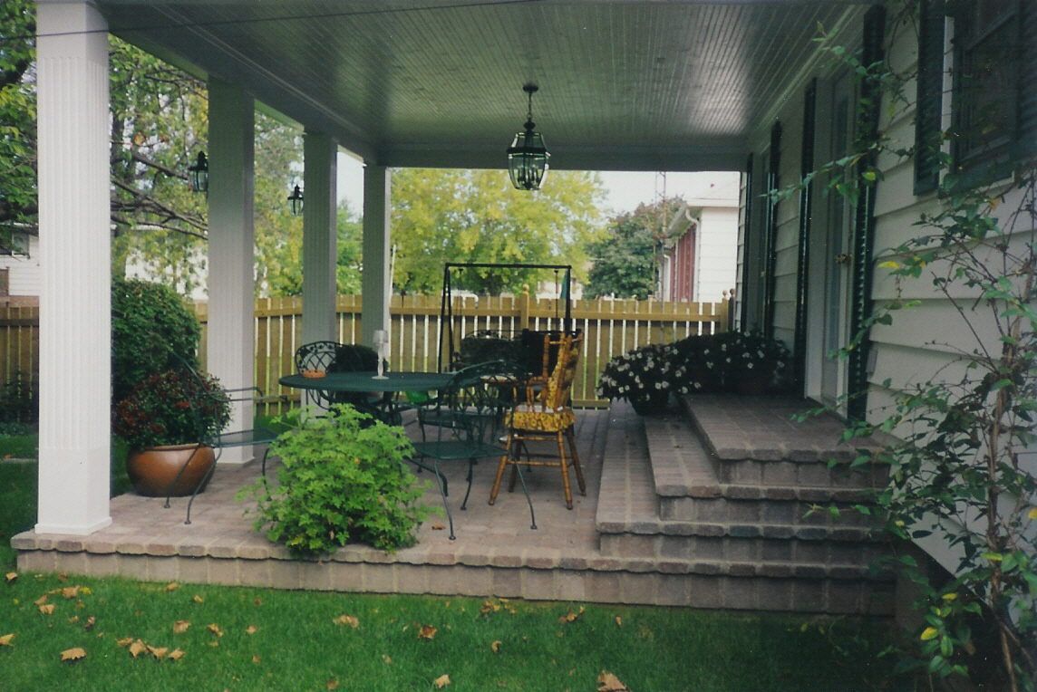 A covered backyard patio with a stone floor, outdoor table, chairs, and brick steps leading to a house entrance.
