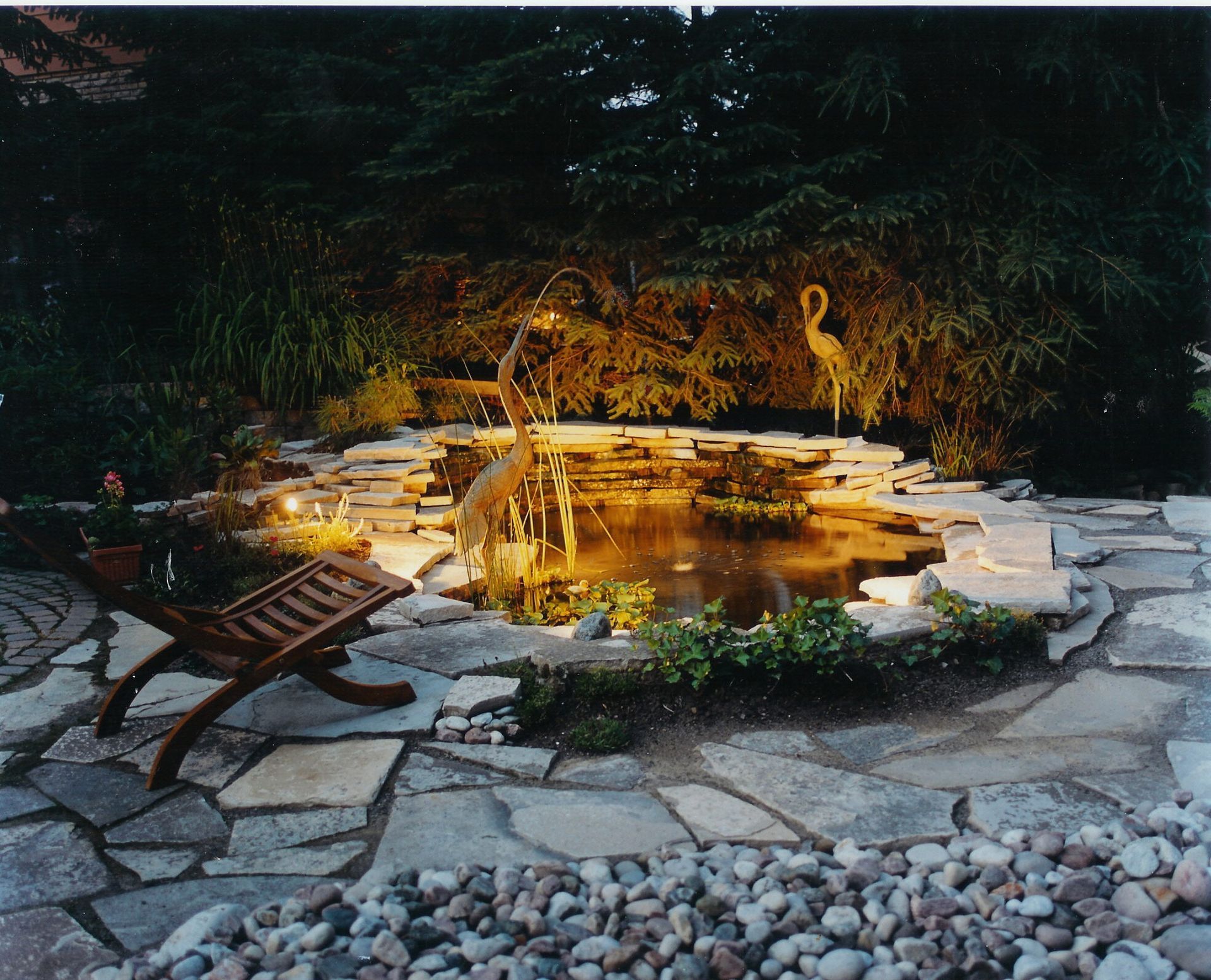An illuminated garden pond at night, featuring tiered rock walls, decorative crane statues, and a wooden lounge chair.