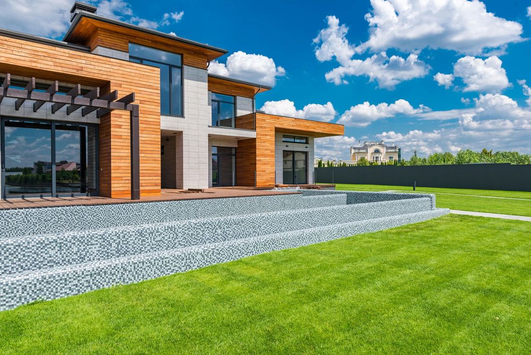 Modern two-story house with wood-paneled exterior, glass windows, and a paved stone patio leading to a lush green lawn.
