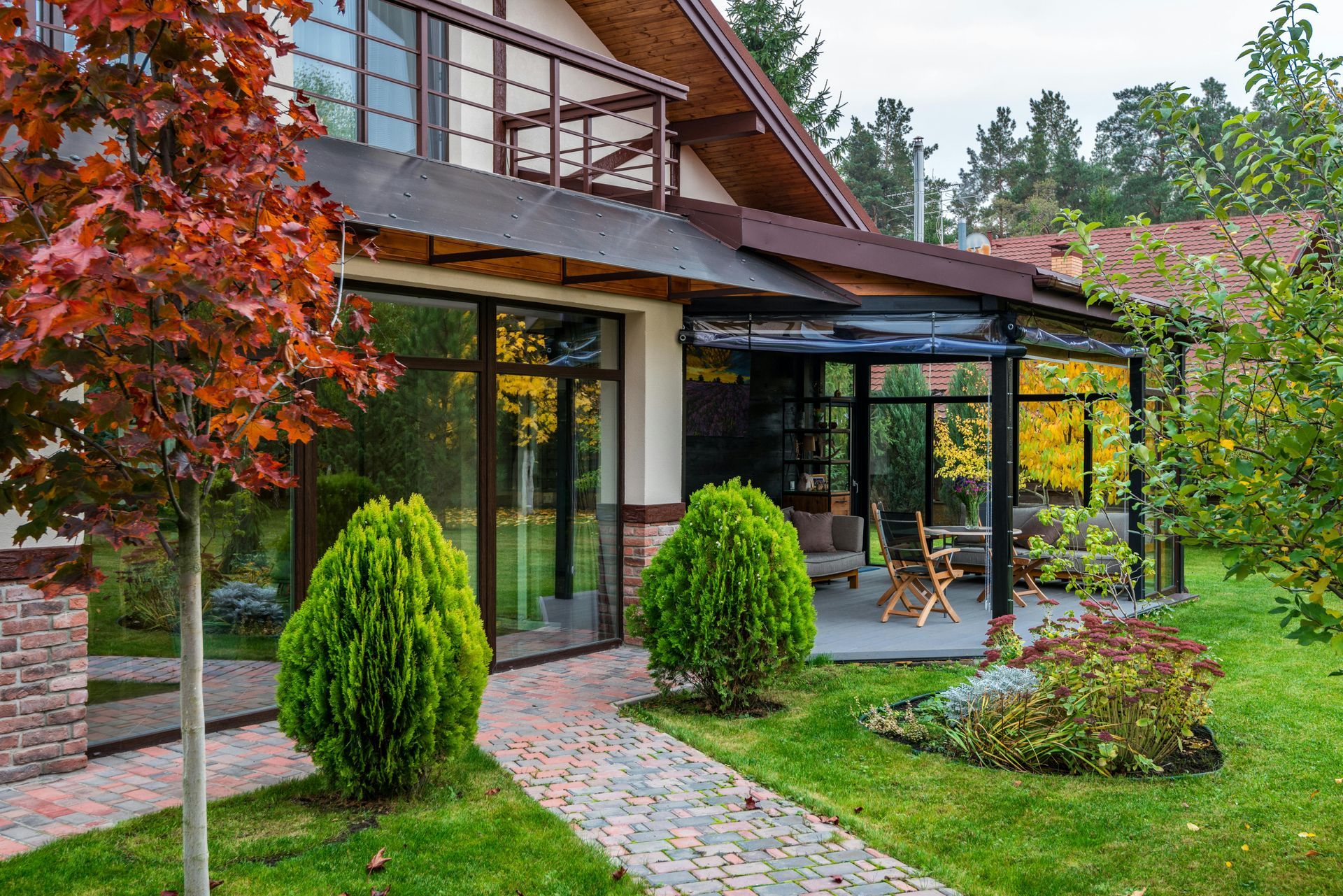 A modern house with a glass-enclosed patio, brick path, and landscaped trees in a yard during autumn.
