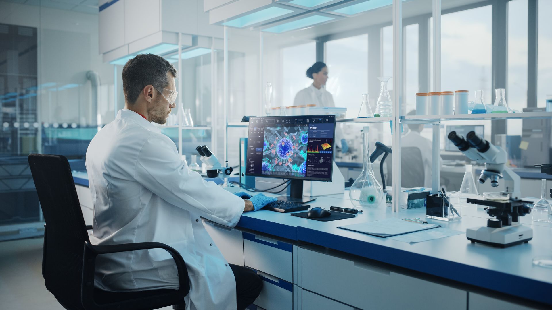 Scientist working at a lab desk with microscope, computer monitors, and test equipment in a bright modern laboratory