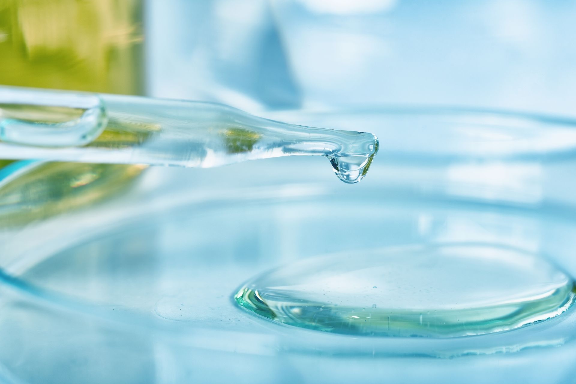 Glass pipette dripping clear liquid into a shallow pool of water on a light blue background