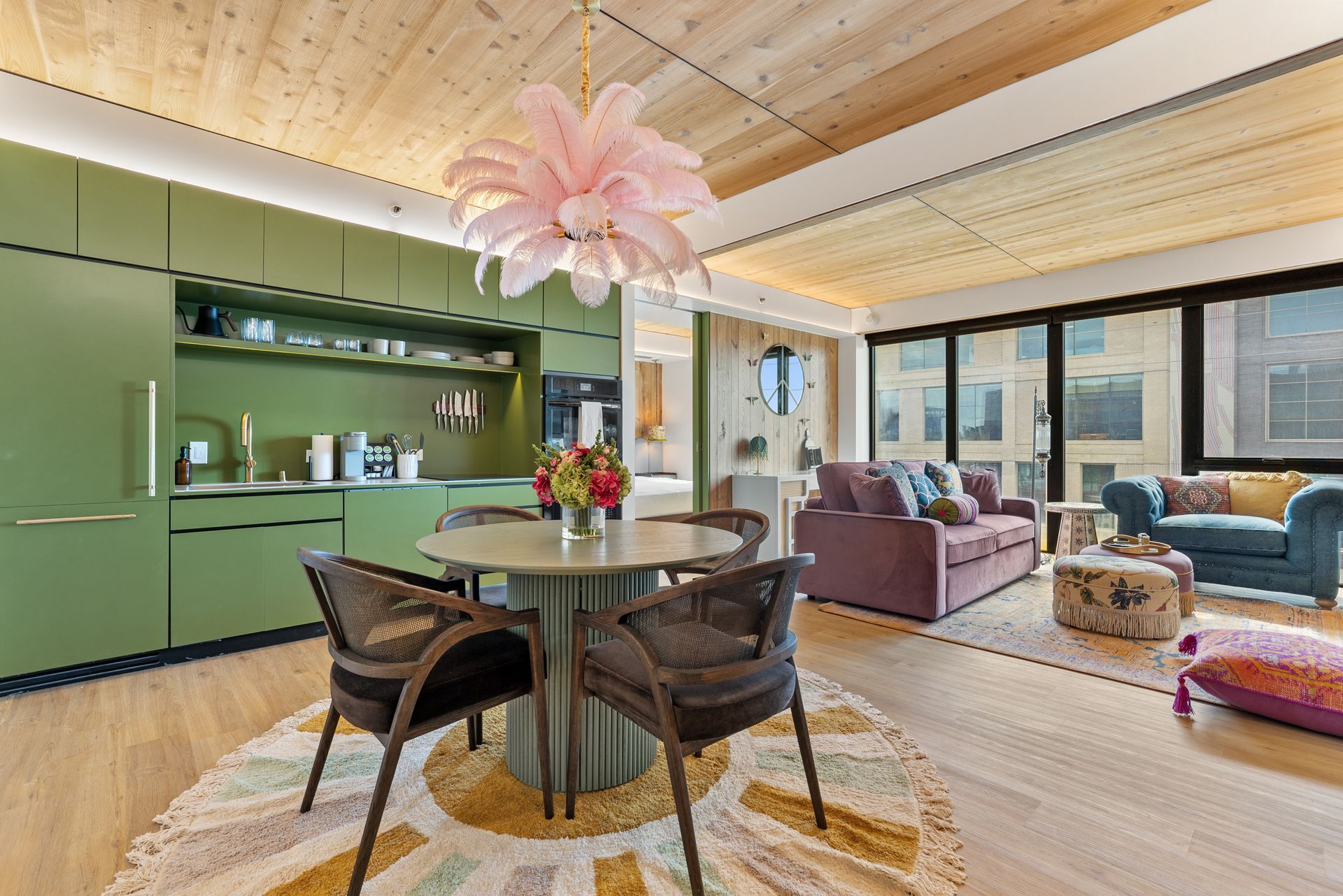 Modern open-plan kitchen and living room with green cabinets, round table, pink pendant light, and large windows