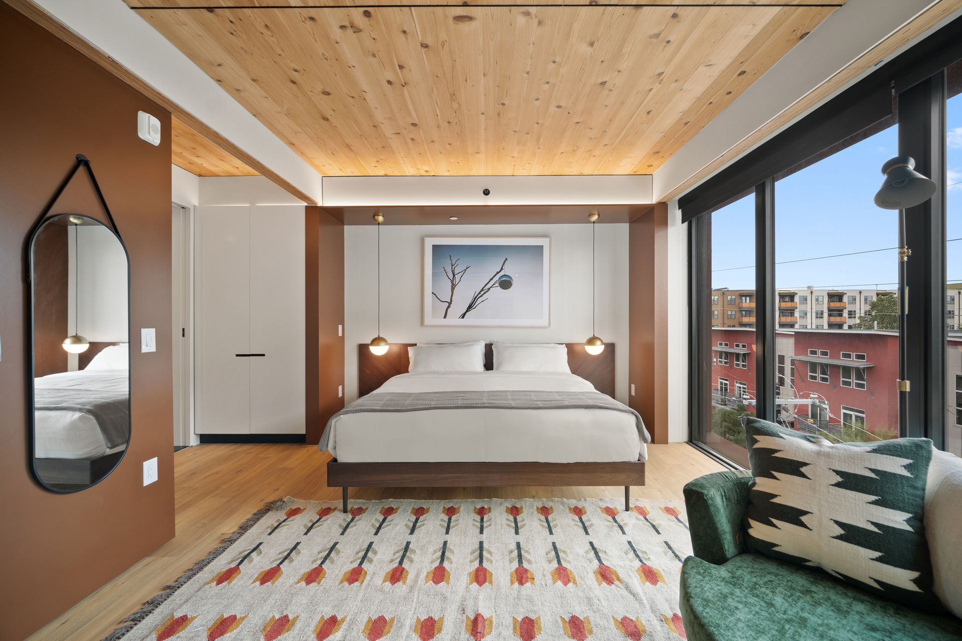 Modern bedroom with bed, wood ceiling, colorful rug, and large city-view window