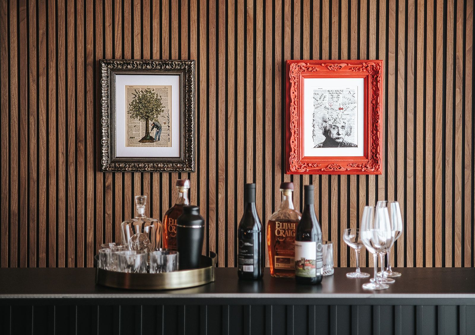 Bar with liquor bottles and glasses on a black counter beneath two framed artworks on a wood slat wall