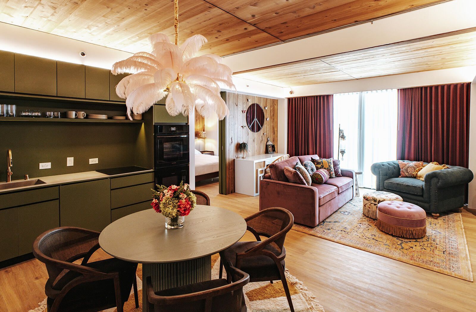 Modern open-plan kitchen and living room with green cabinets, round table, pink pendant light, and large windows