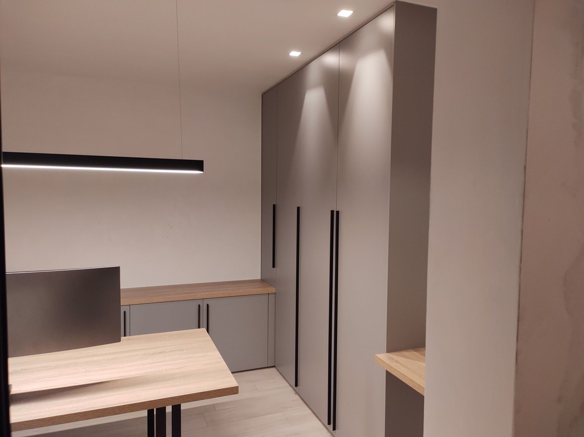 Een moderne kantoorruimte met lichte houten bureaus en grijze kasten. Een lange zwarte lamp hangt boven de bureaus.