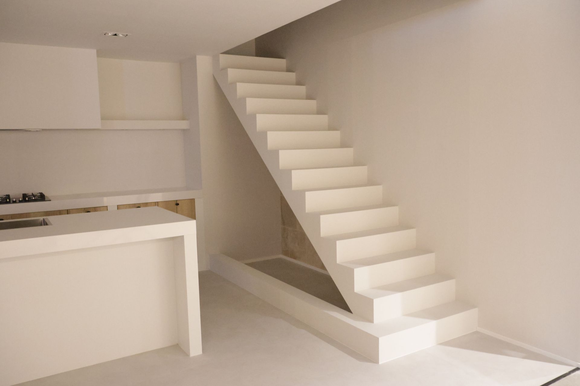 Witte trap tegen een witte muur, naast een aanrecht en apparatuur. De setting is modern en minimalistisch.