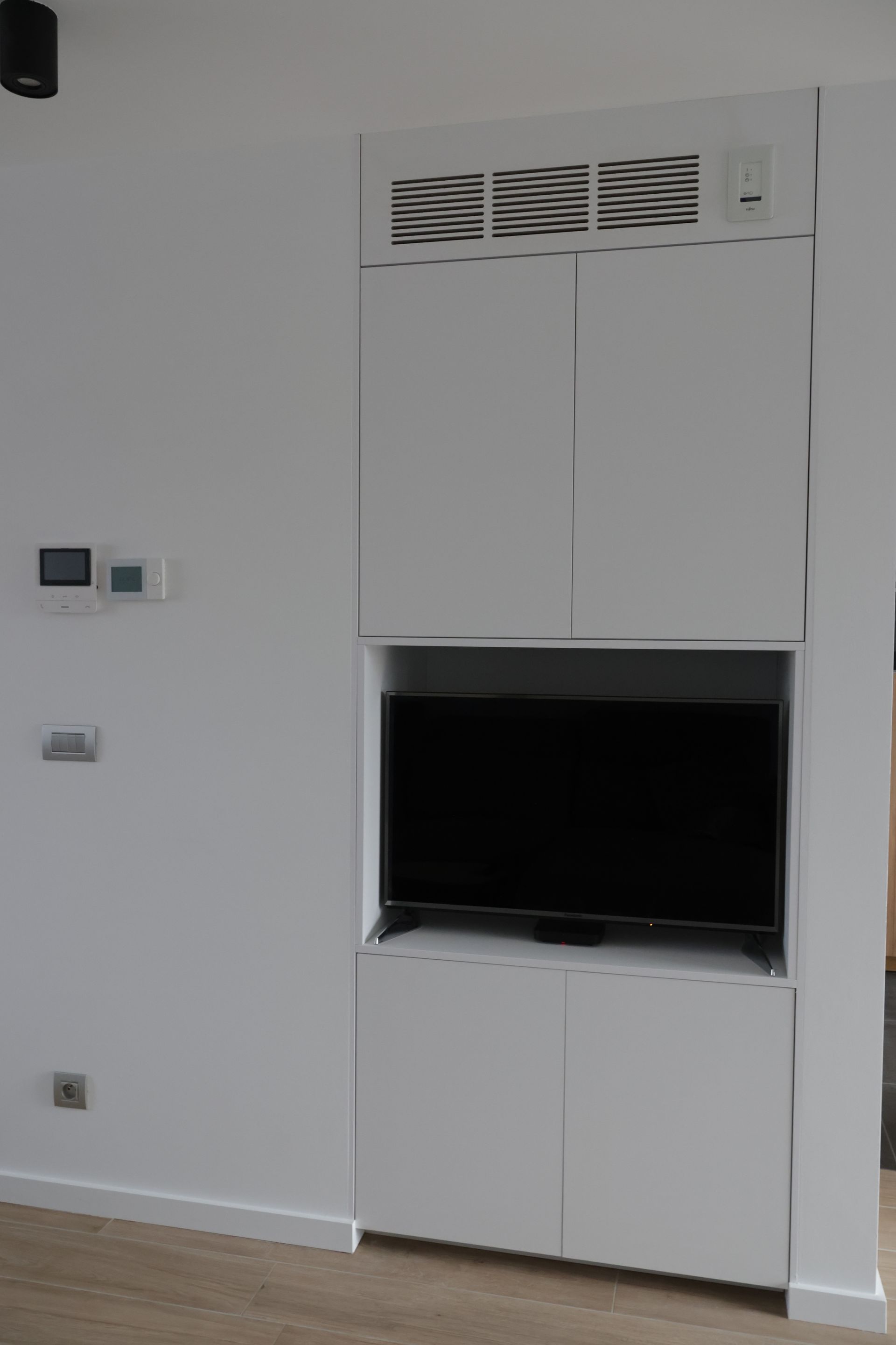 Ingebouwde witte kast met tv, ventilatie en opbergruimte. Gemonteerd aan een witte muur met thermostaat.