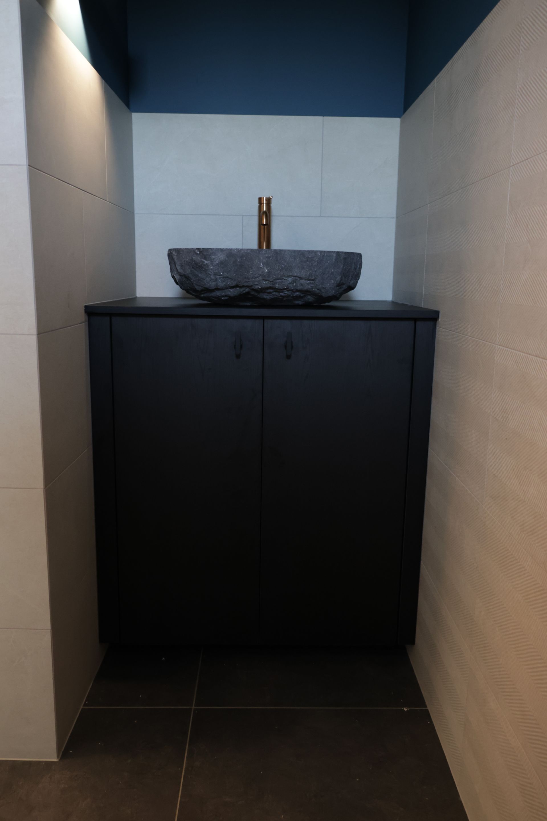 Donkere, moderne badkamermeubel met een stenen wasbak en bronzen kraan. De kast is zwart en staat mooi tegen de grijze muren.