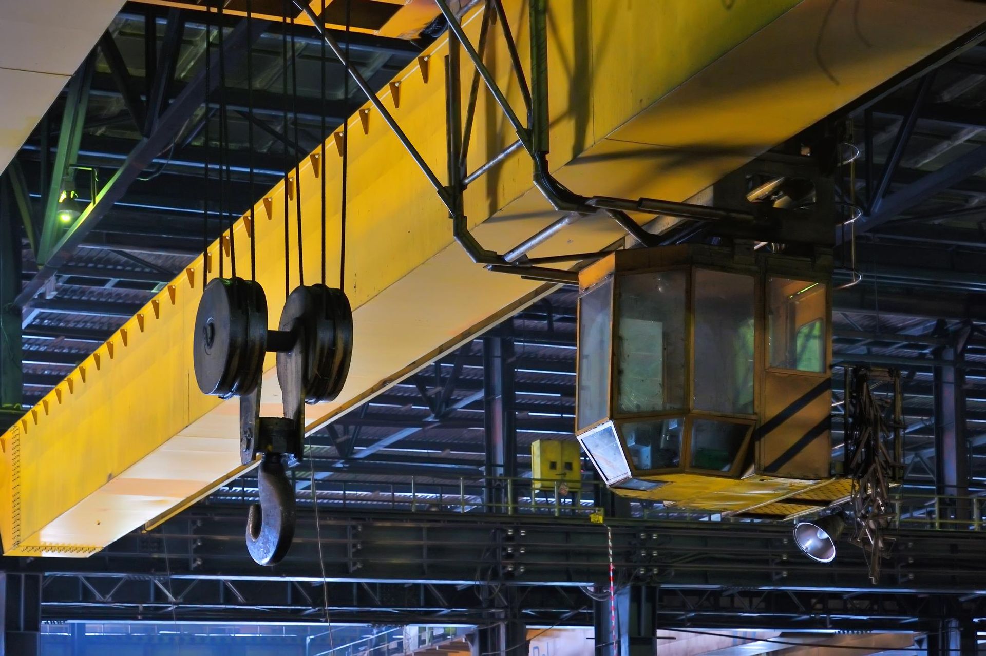 Yellow overhead crane in a factory setting. Hook and pulley system visible with a control cabin.