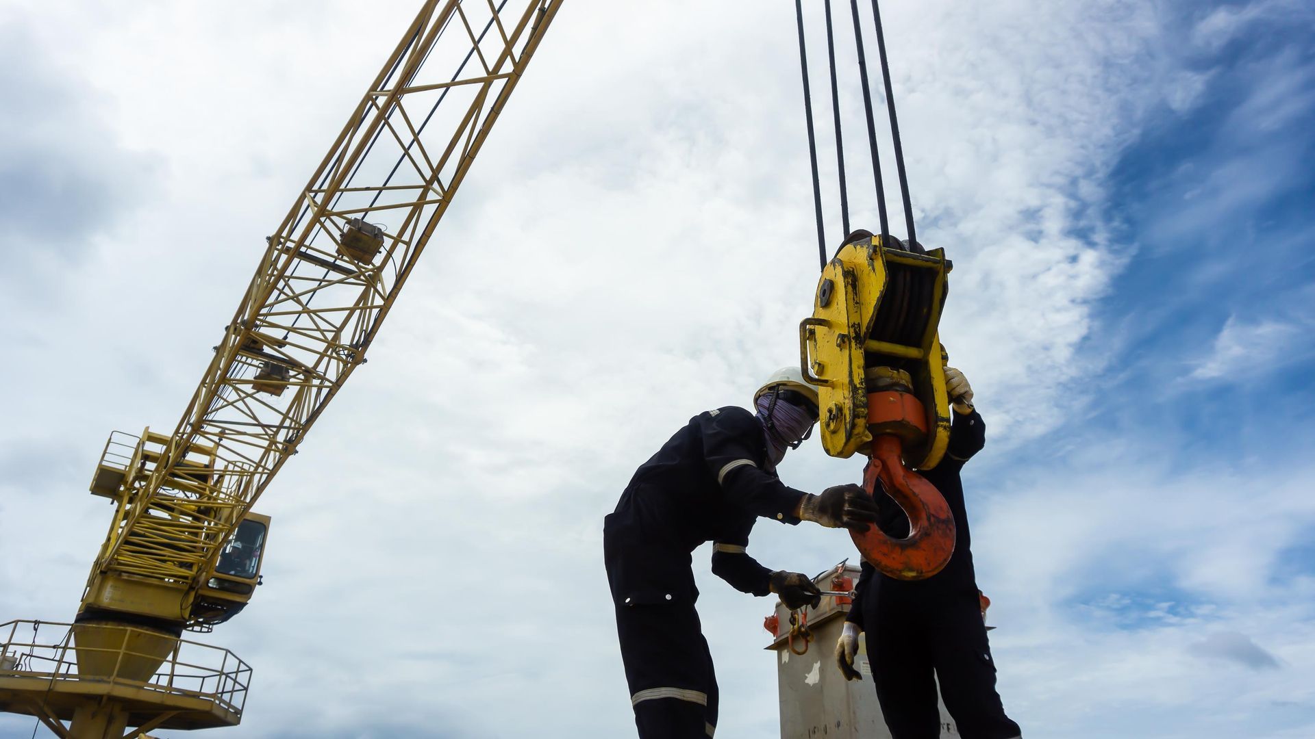 Worker connecting equipment to crane hook against a cloudy sky.