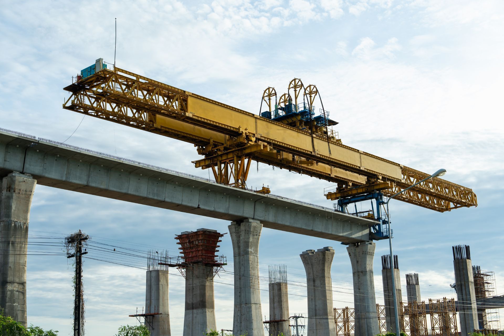 A yellow girder bridge construction crane installing a concrete beam on elevated support columns, under a cloudy sky.