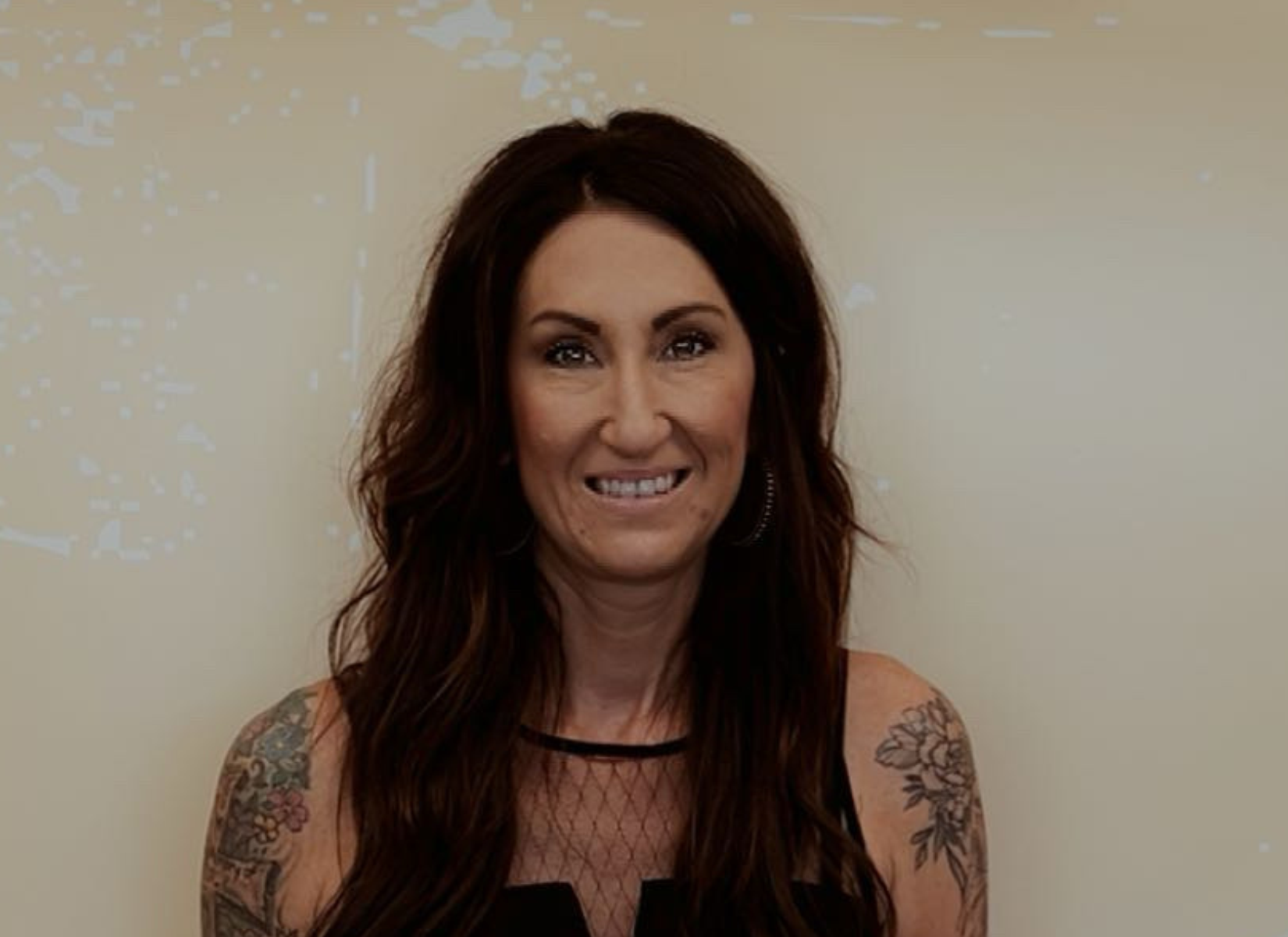 Woman with long dark hair, smiling, tattoos on arms, in front of a beige wall.