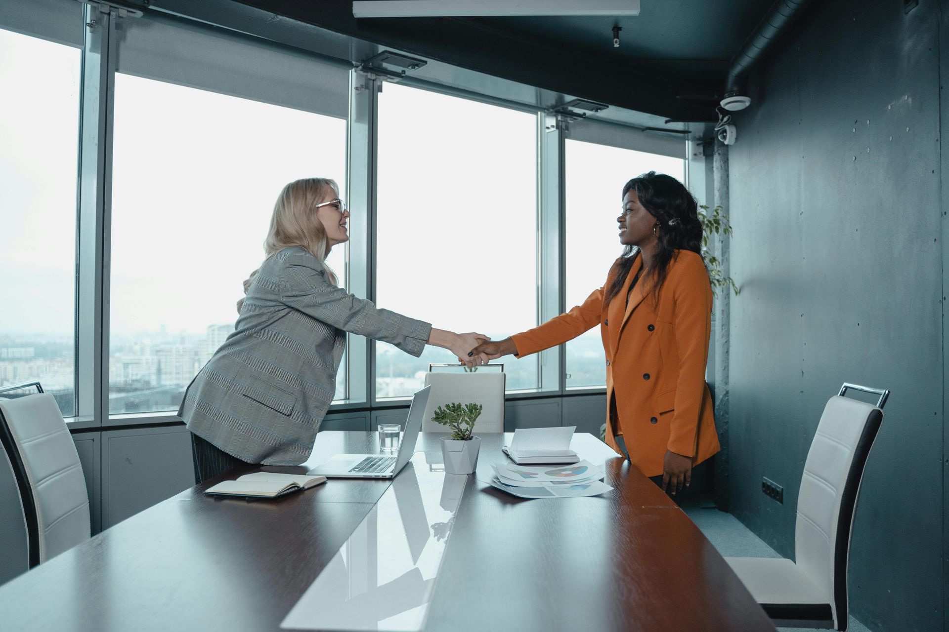 Two women shaking hands in a modern office with city views, celebrating a deal.