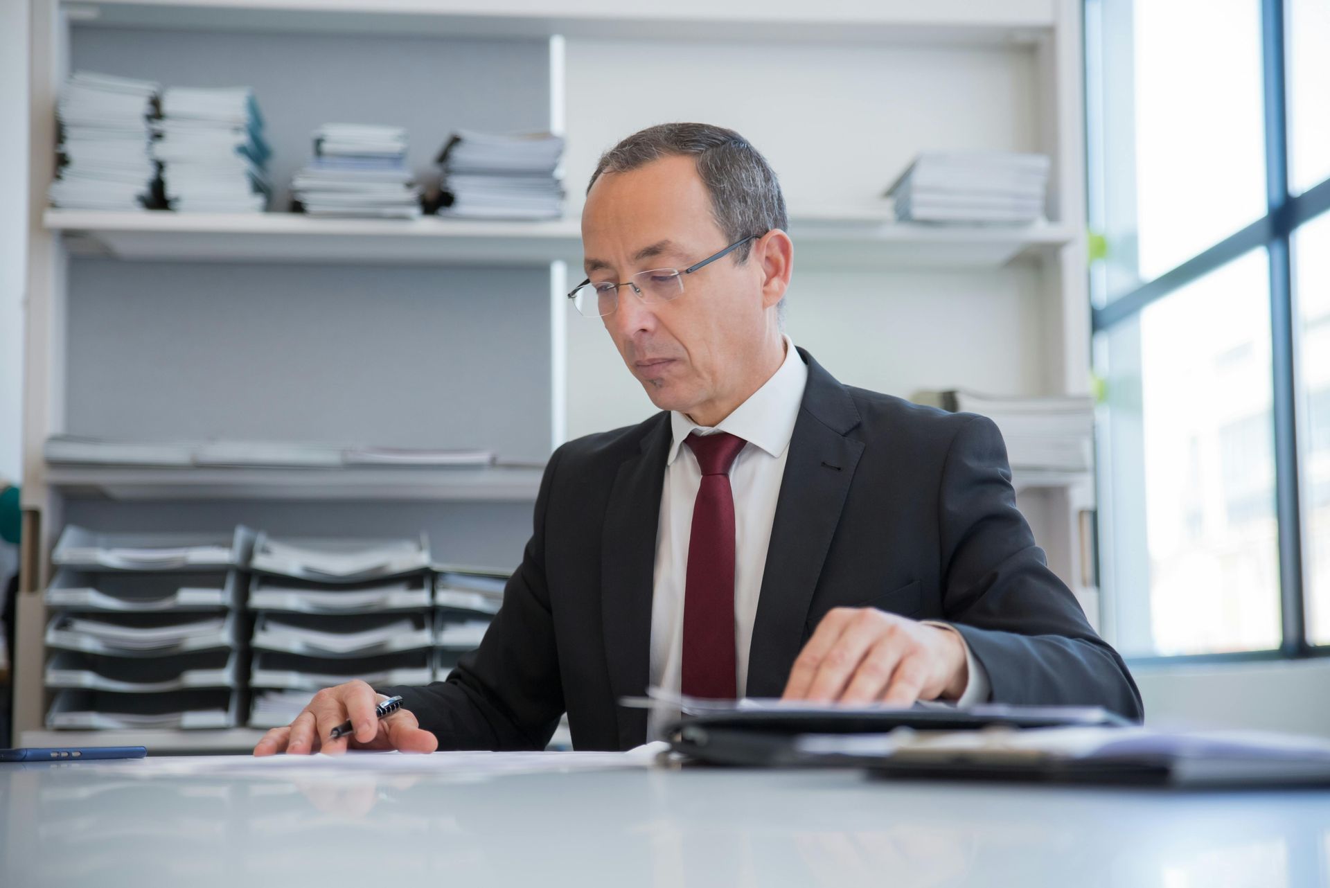 Man in suit at desk, reading documents in office setting.