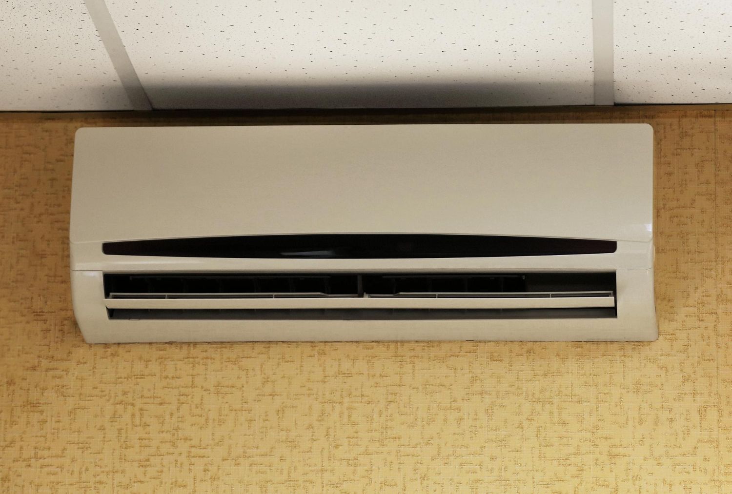 A white, wall-mounted air conditioning unit is positioned below a white drop ceiling against a tan, textured wall.