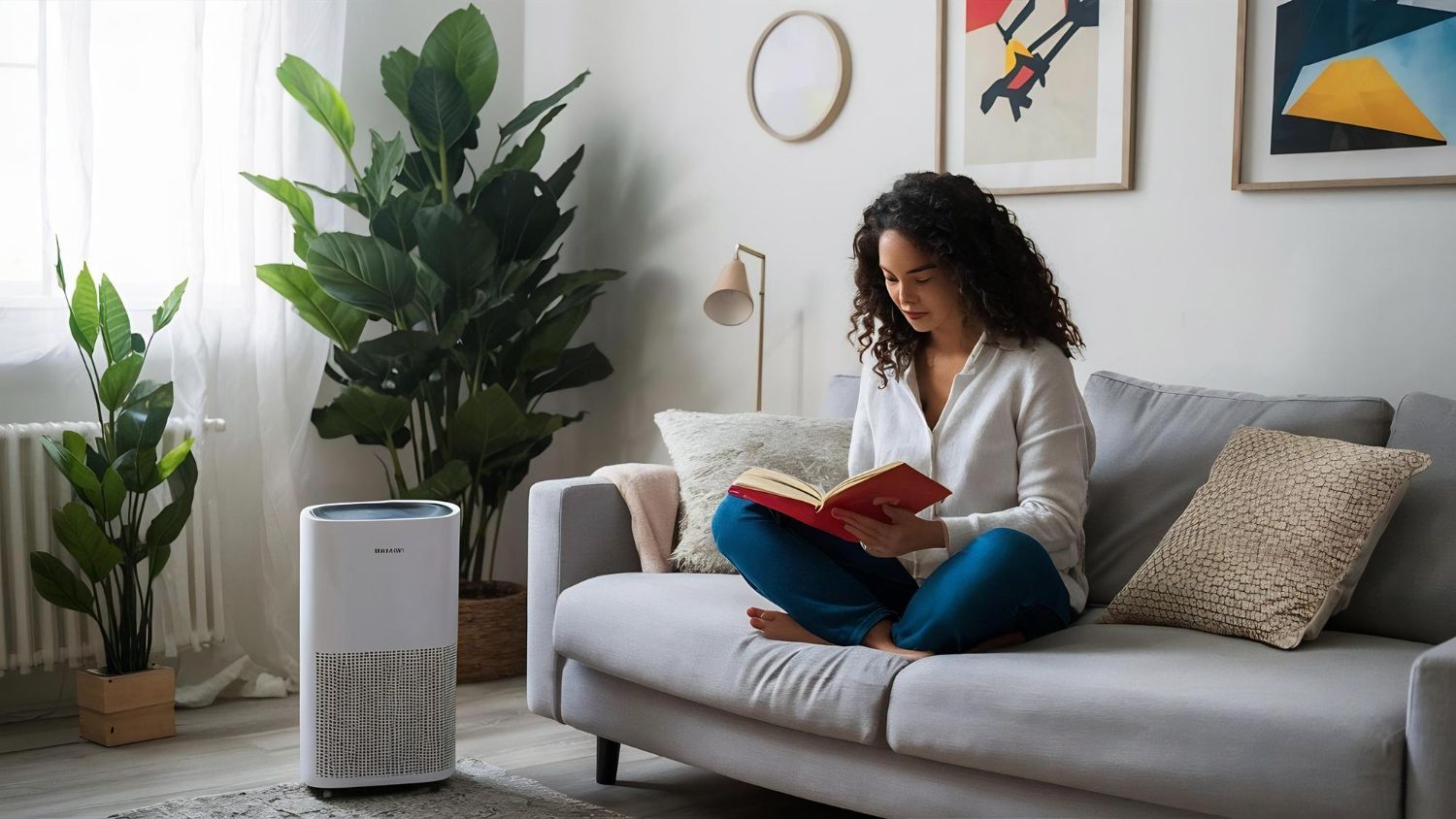 A person sitting on a sofa in a living room, reading a book next to a white air purifier and a large potted plant.