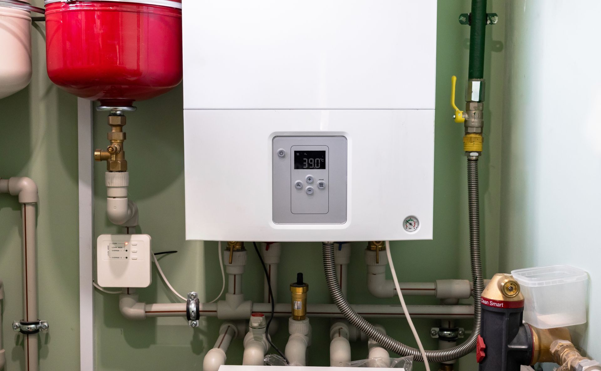 White wall-mounted boiler with pipes and valves in a utility room, next to a red expansion tank.