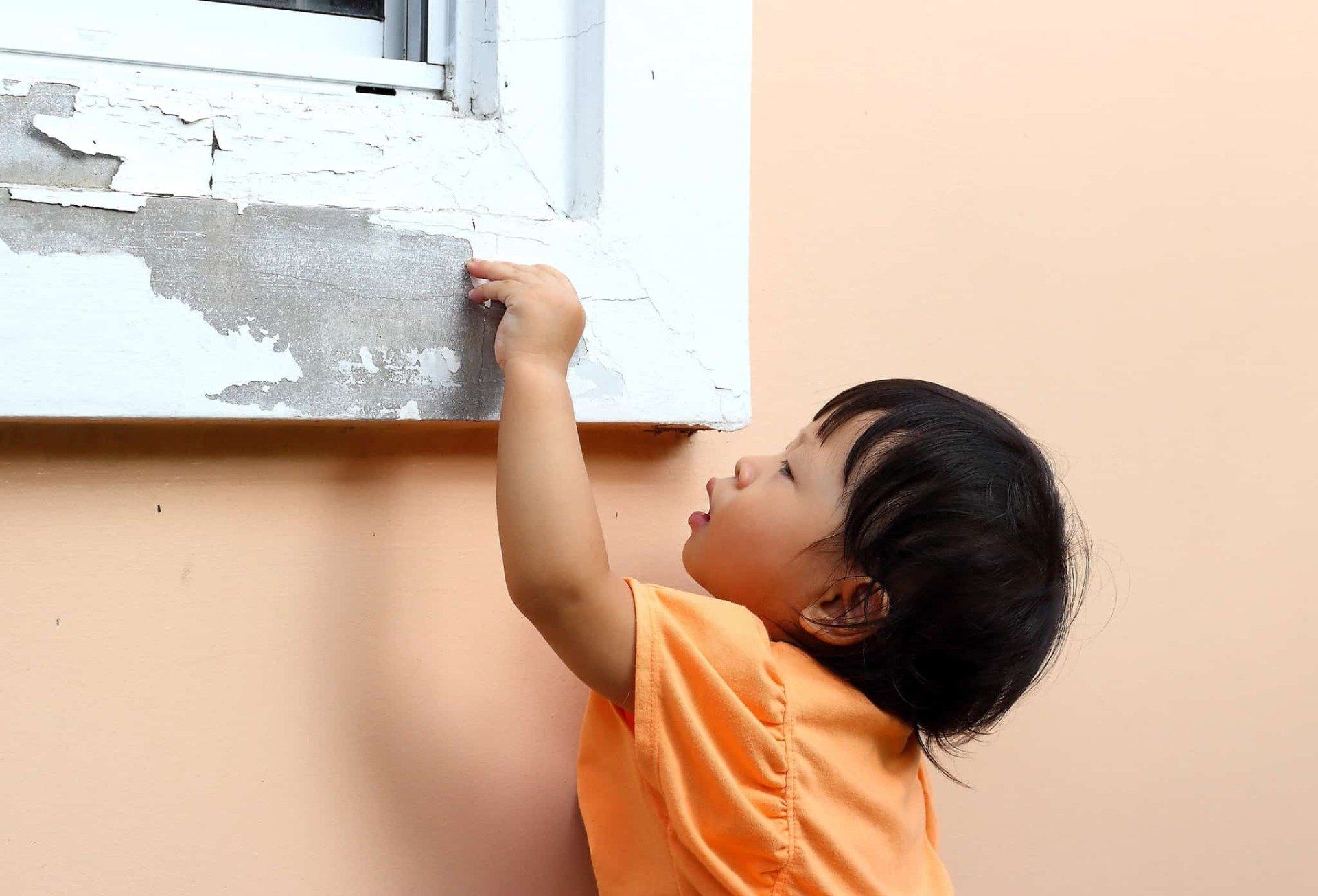 Child in an orange shirt reaching up to touch peeling paint on a window frame