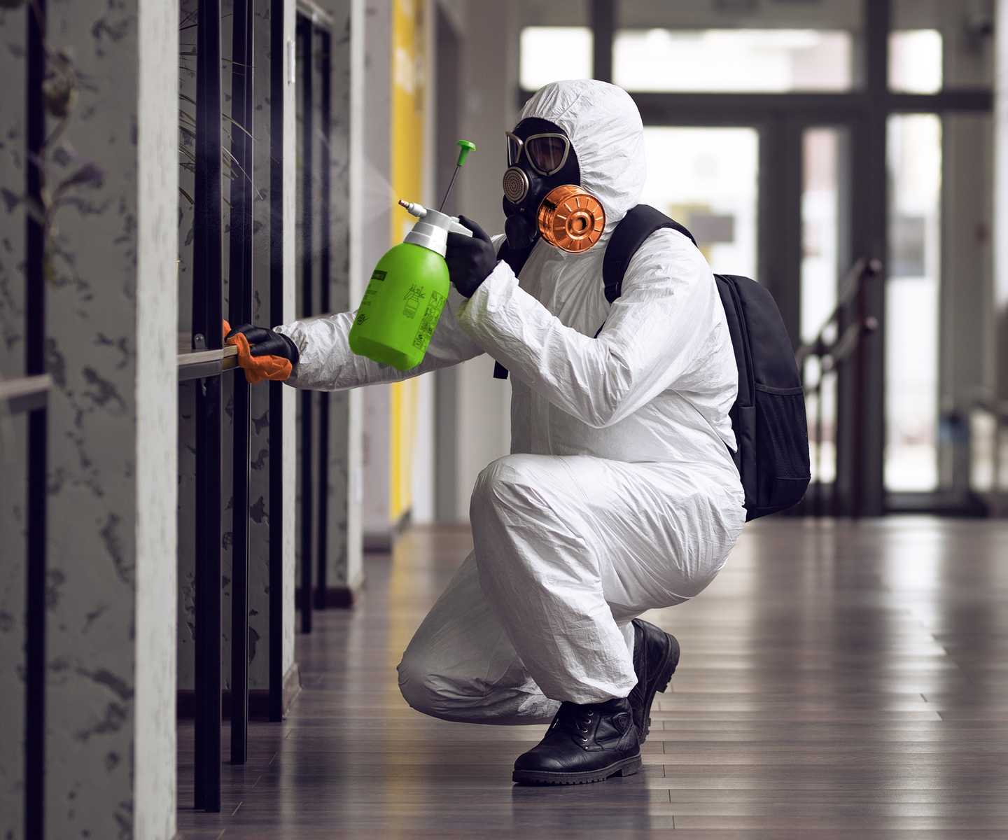 Worker in white protective suit and gas mask spraying disinfectant in a hallway