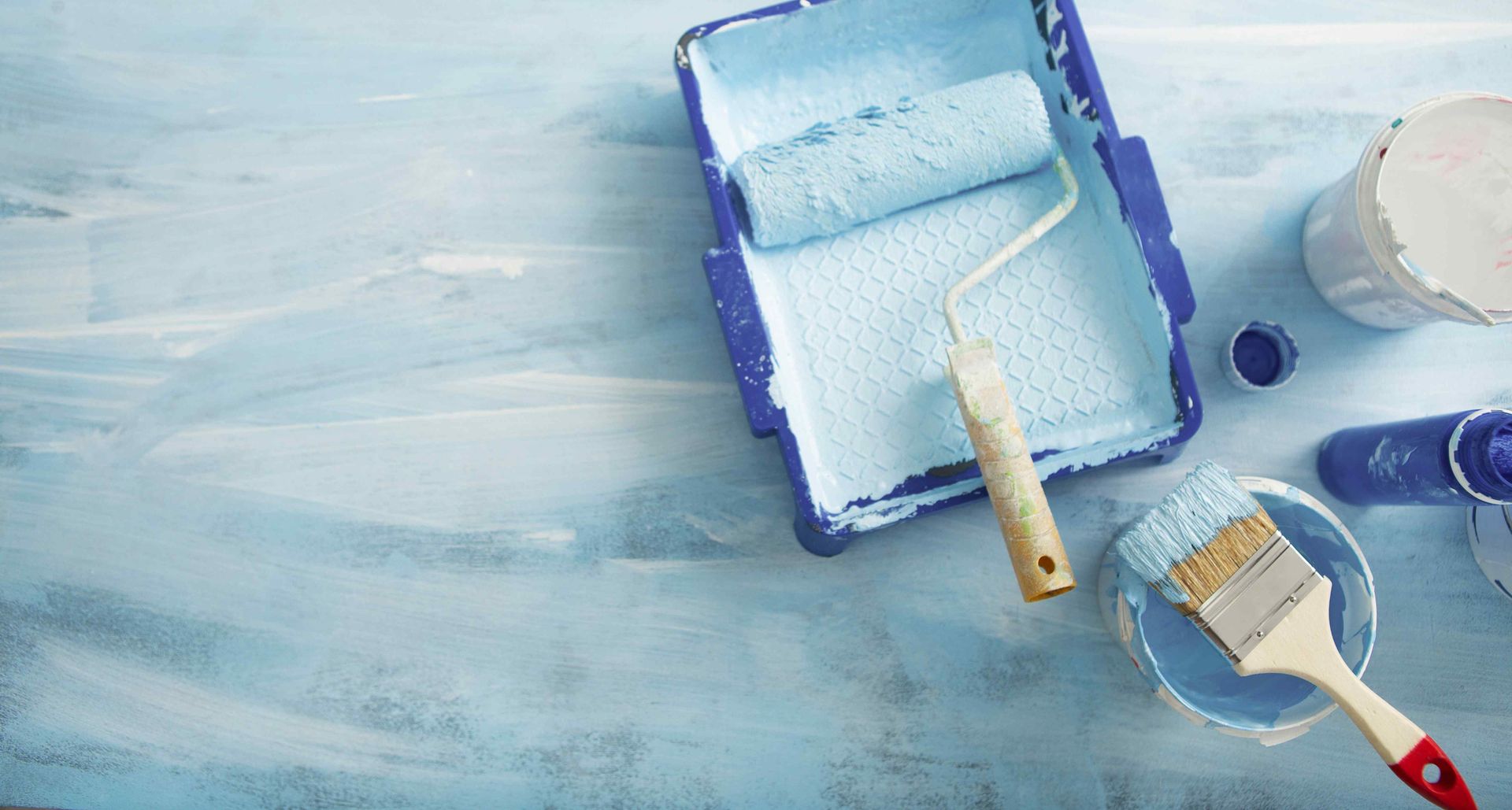 Blue paint supplies on a blue painted surface: roller, tray, brush, and paint cans.