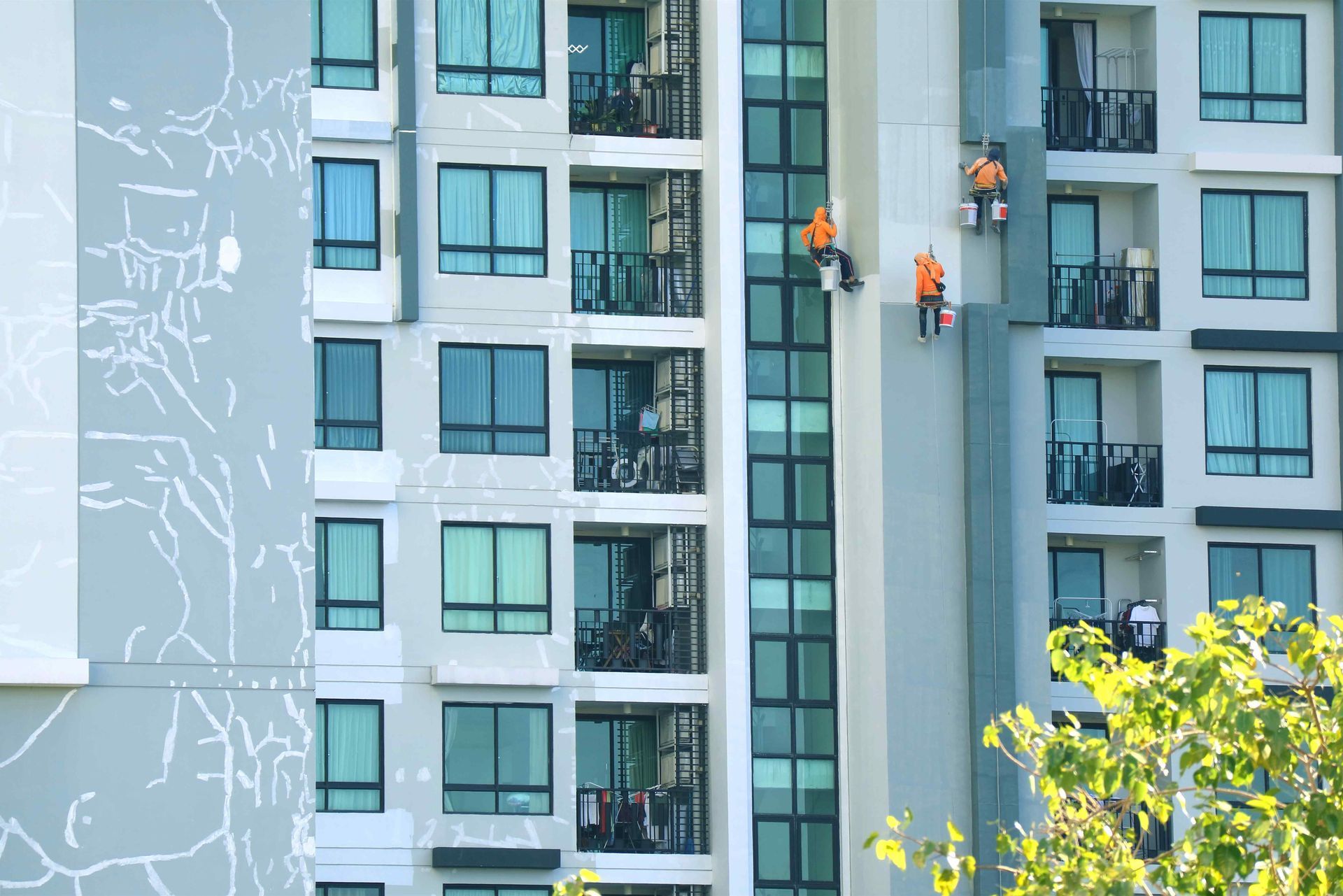 Workers on a high-rise building, painting the facade. Orange safety harnesses and gray exterior.