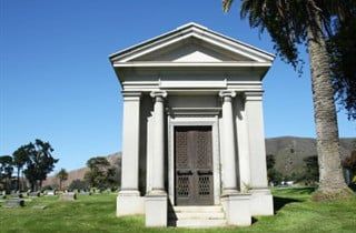 Small white mausoleum with columns in a grassy cemetery under a clear blue sky