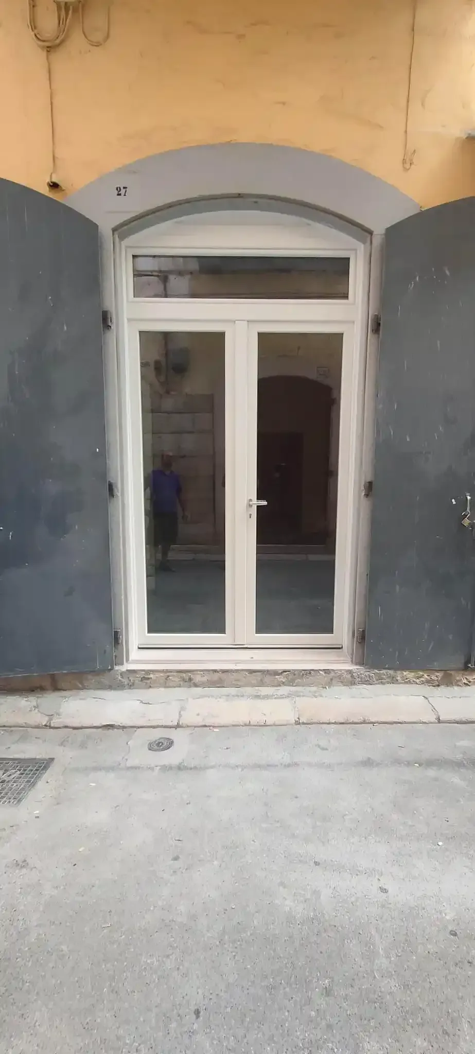 Porte in vetro con cornice bianca inserite in un portale grigio-bluastro sotto un arco giallo. La porta riflette un interno.