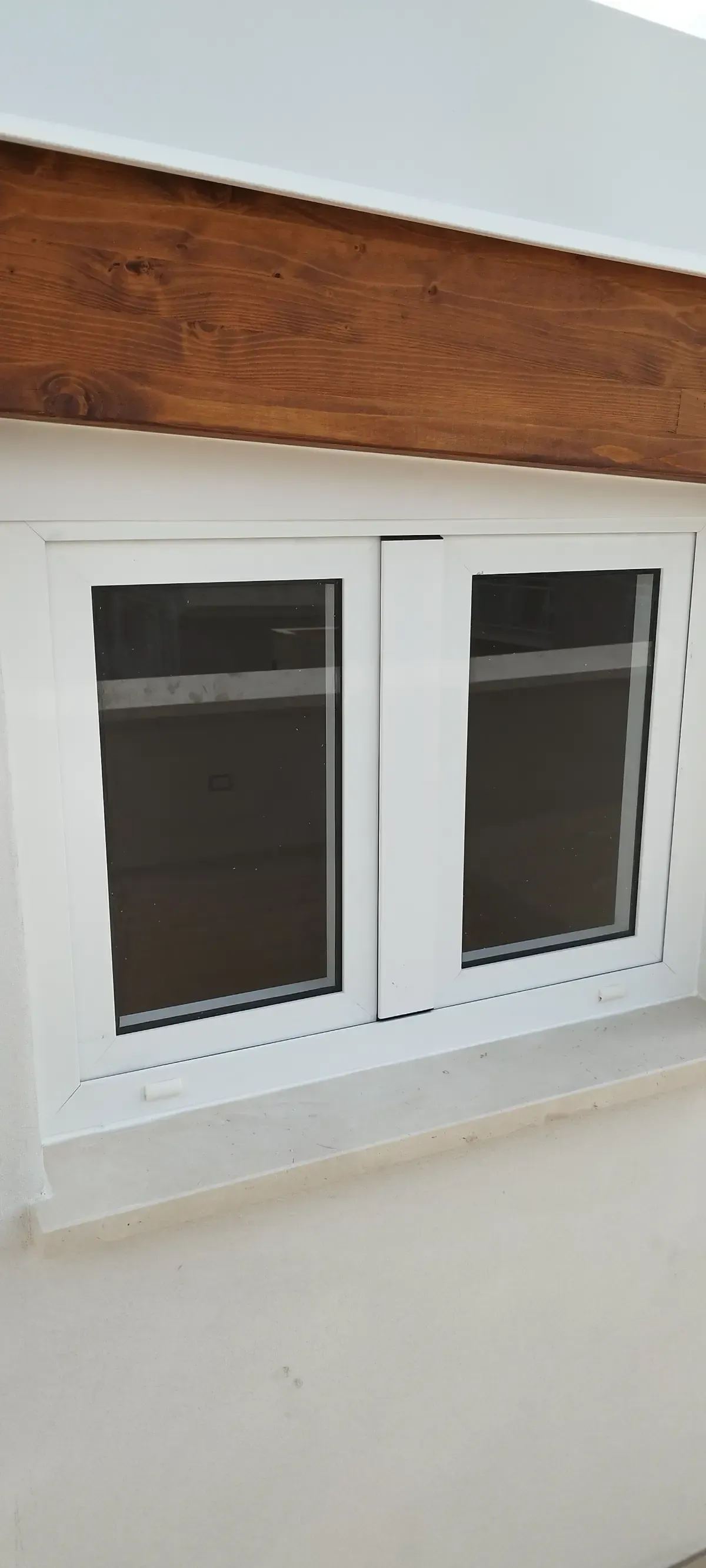 Finestra con cornice bianca e due vetri, incastonata in una parete bianca sotto una trave di legno marrone.