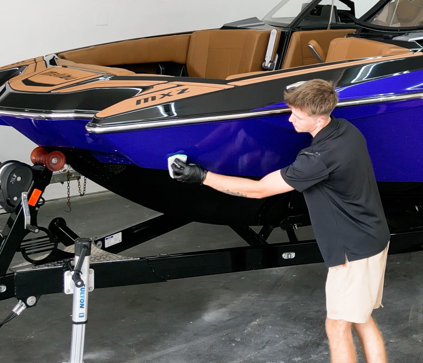 Man wearing gloves cleaning a blue boat on a trailer with a white cleaning pad.