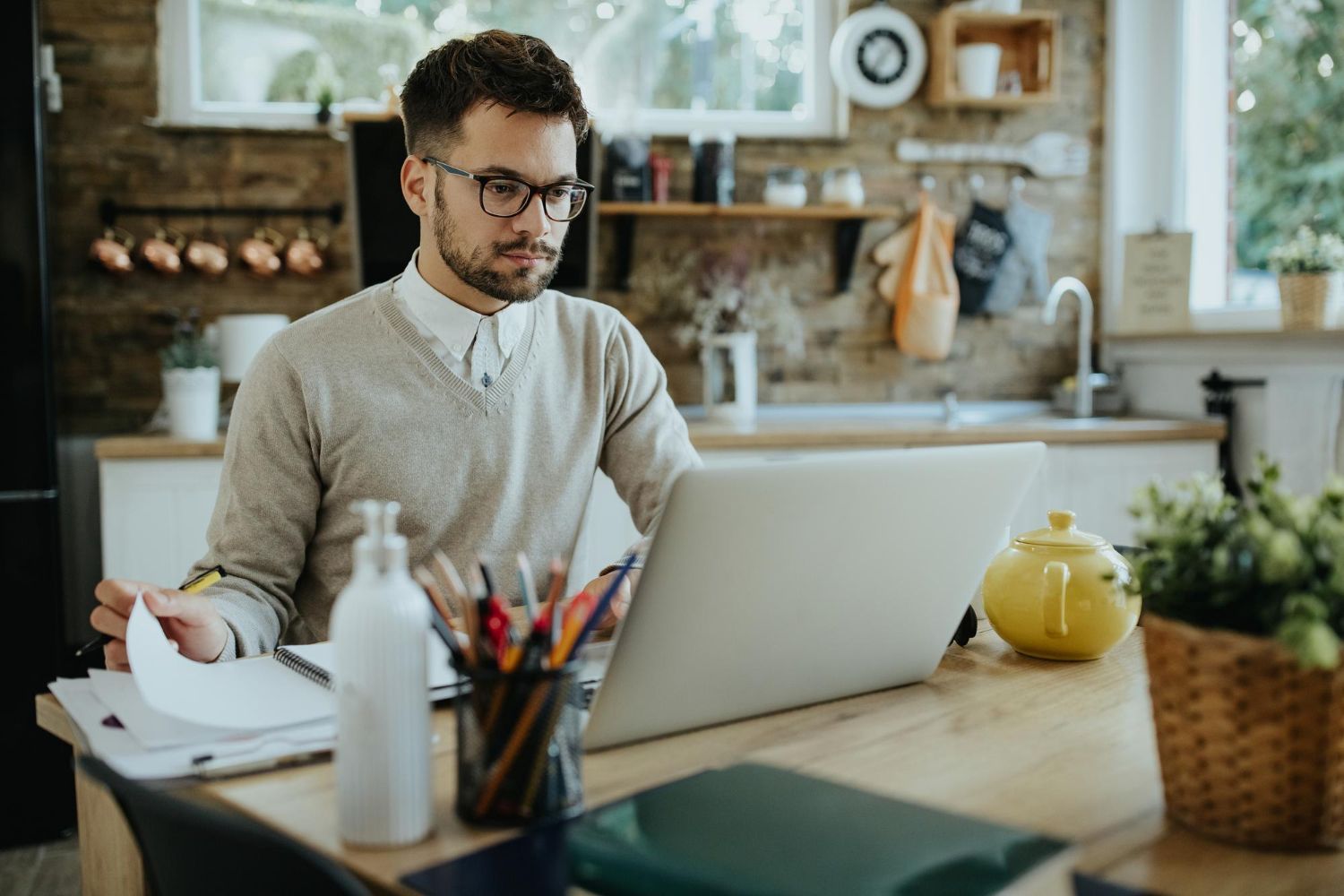 Person working at a laptop in a bright kitchen, surrounded by notebooks, pens, and a teapot.