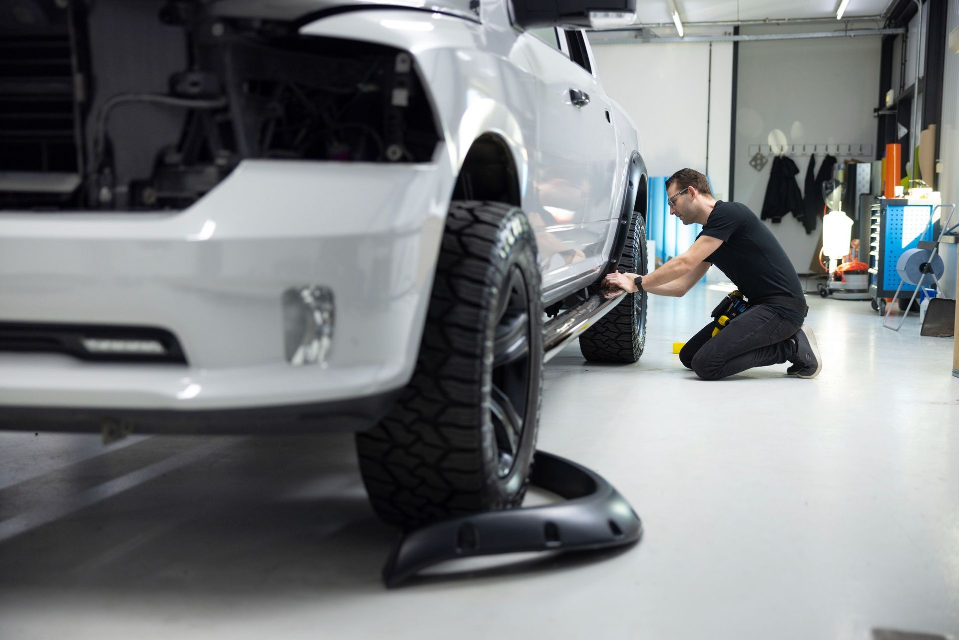Mechanic inspecting a white SUV in a garage, using tools near the front wheel.