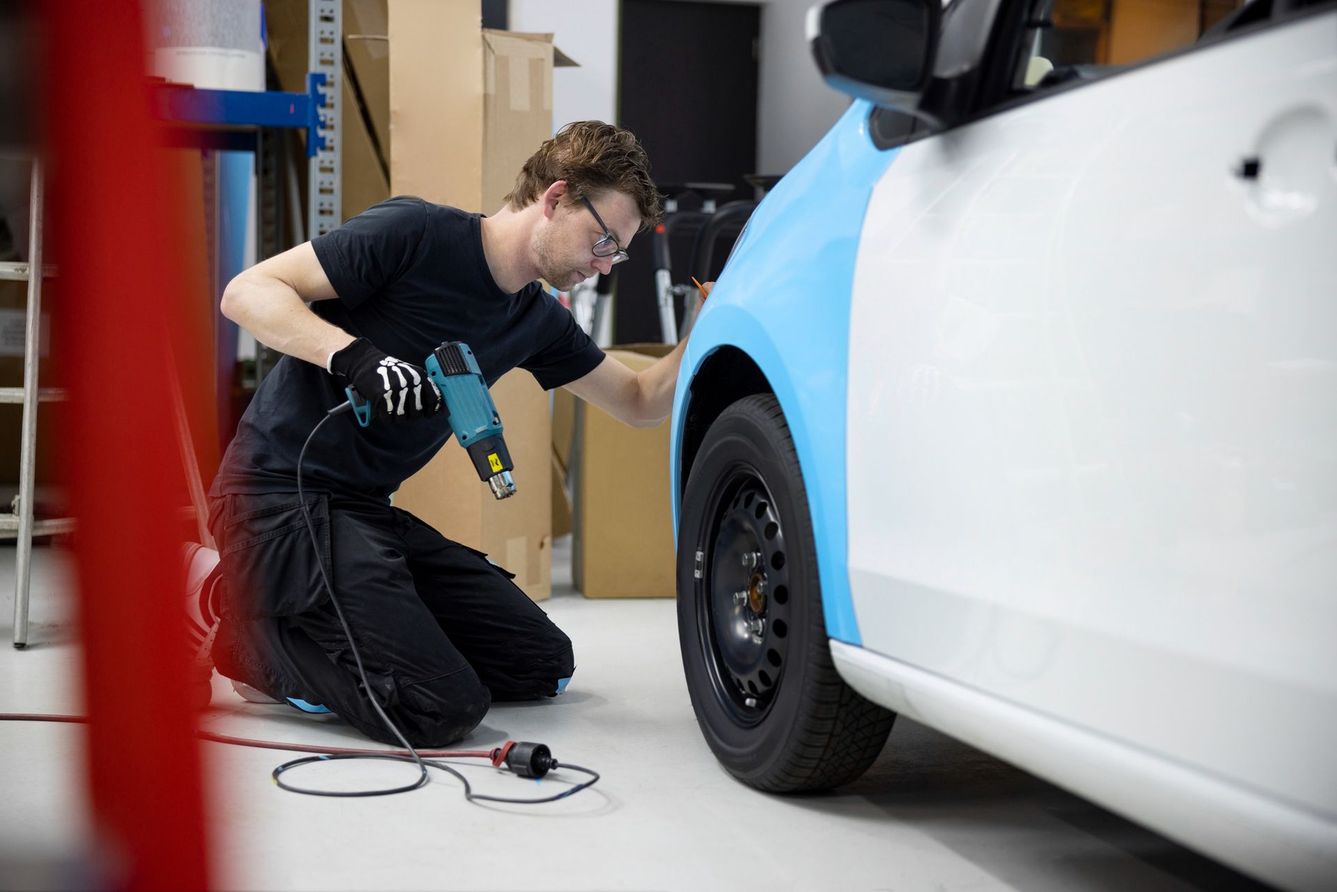 Technician kneels beside a white car, using a drill near the front wheel in a workshop.