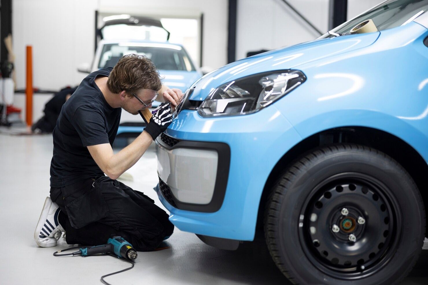 Mechanic kneeling and inspecting the front of a light blue car in a garage