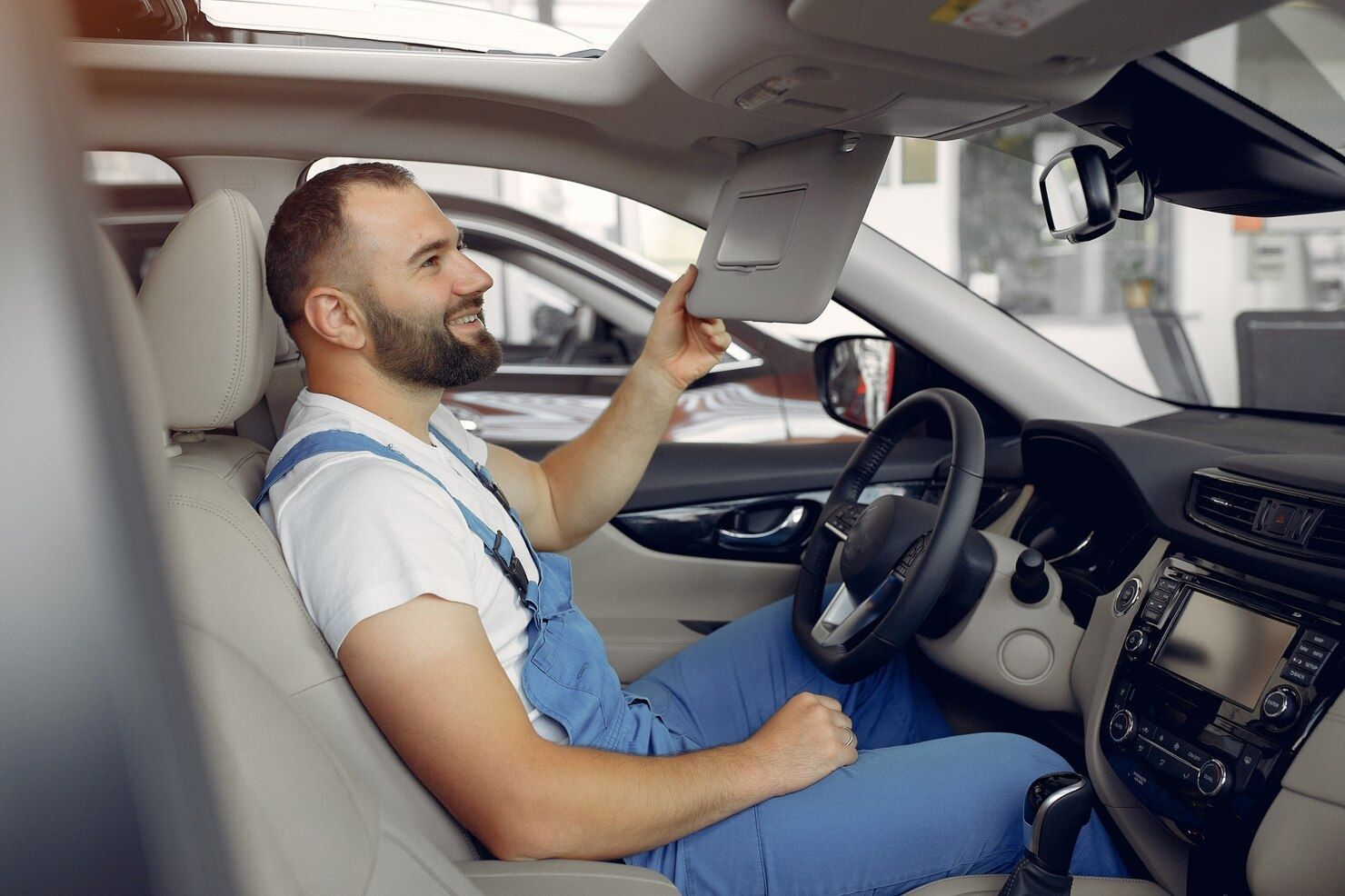 Man in blue overalls sitting in a car showroom, inspecting the roof controls inside a white vehicle.