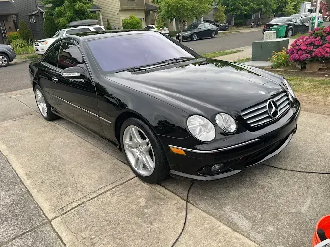 Black Mercedes-Benz CL coupe parked on a concrete driveway, next to a street with other parked cars and houses.