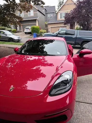 Red Porsche sports car parked in a residential neighborhood, door open. Other vehicles and houses in the background.