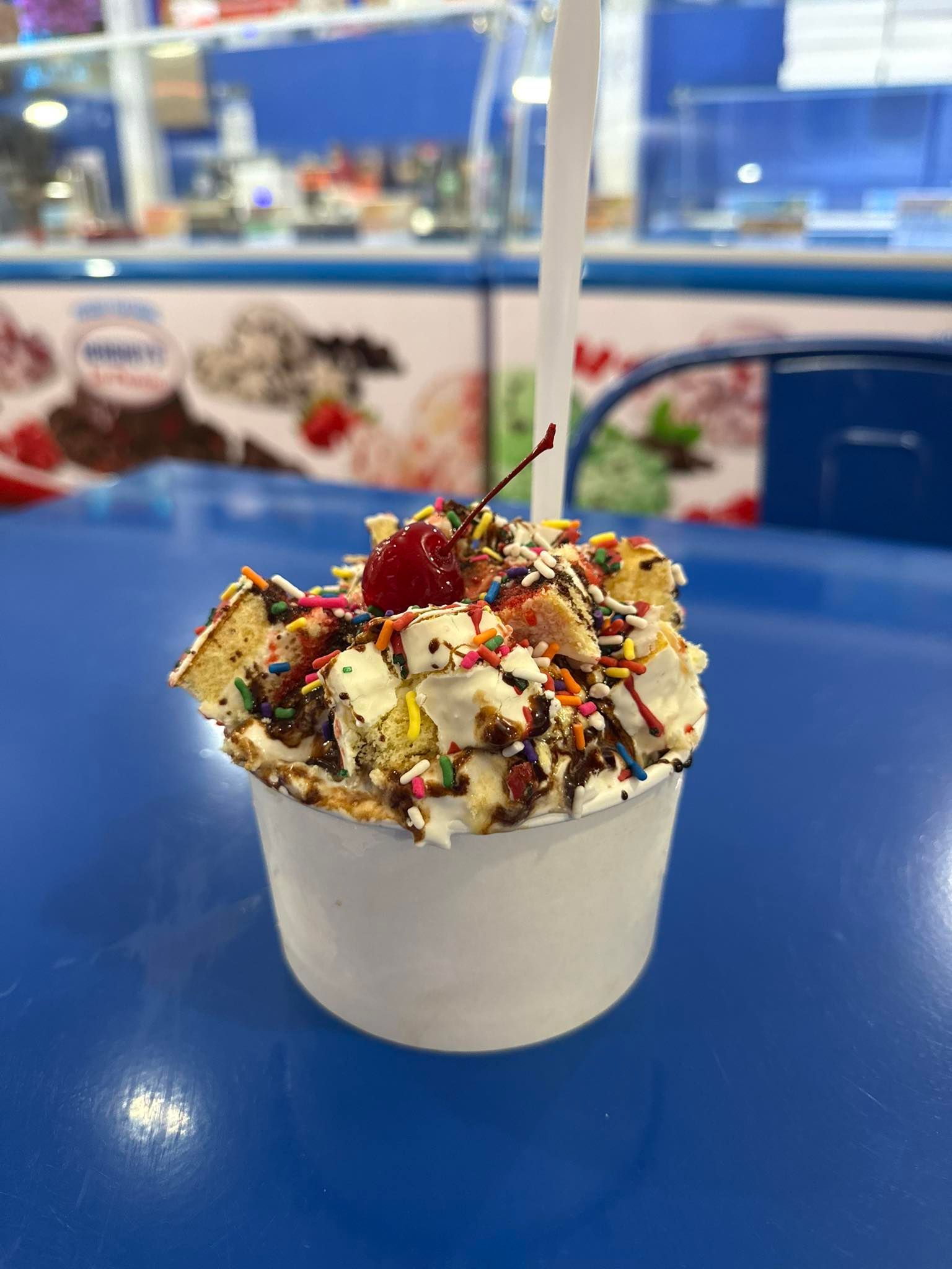 A white cup of ice cream topped with whipped cream, sprinkles, and a cherry, sitting on a bright blue table.