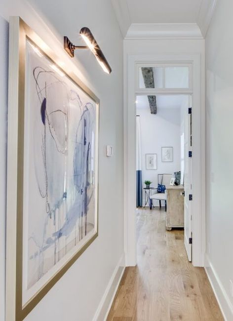 A bright, narrow hallway with light wood floors and a large blue and white abstract painting lit by a modern wall lamp.