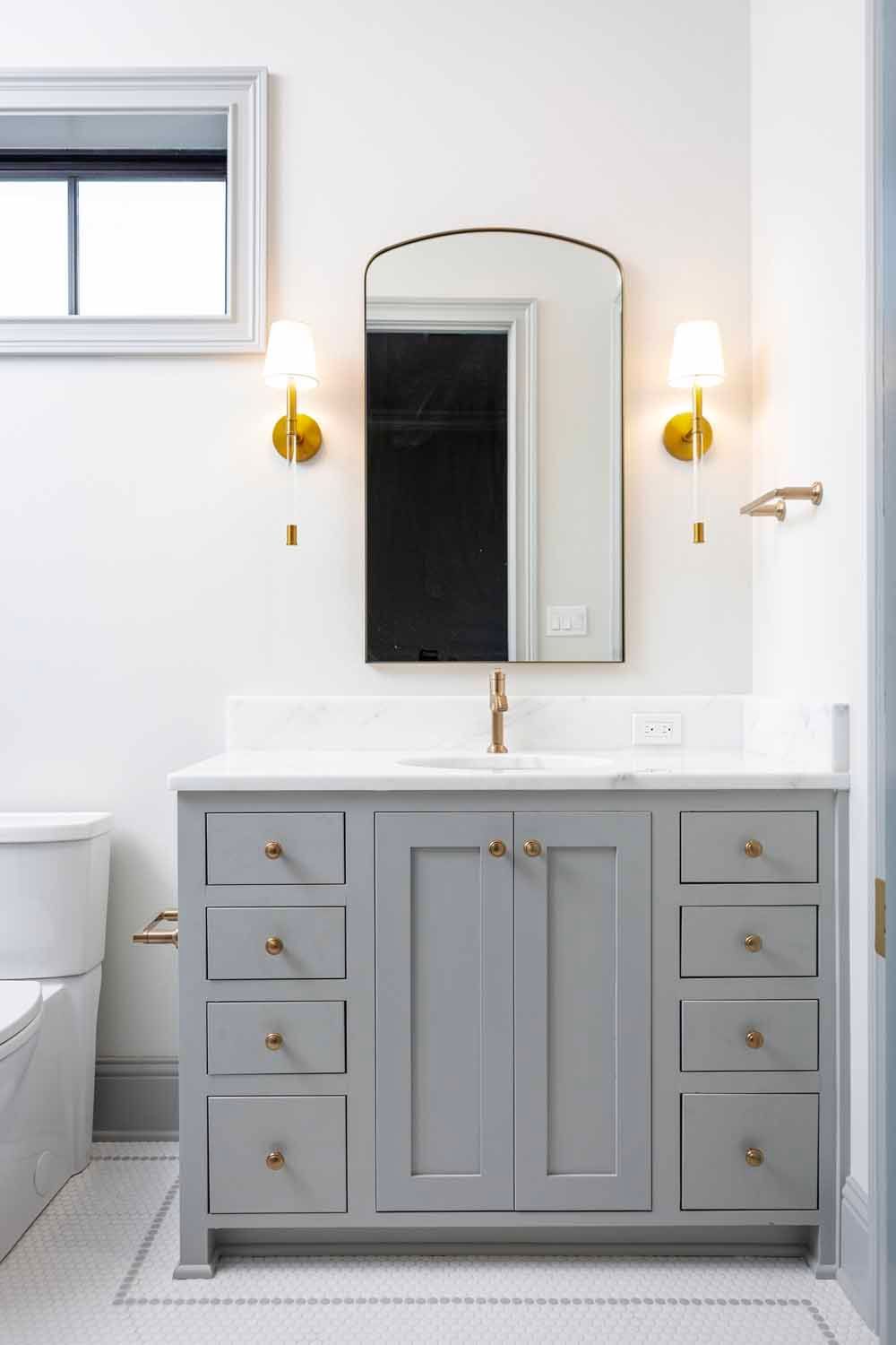 A gray bathroom vanity with gold hardware, topped by a marble counter, brass faucet, and arched mirror between sconces.