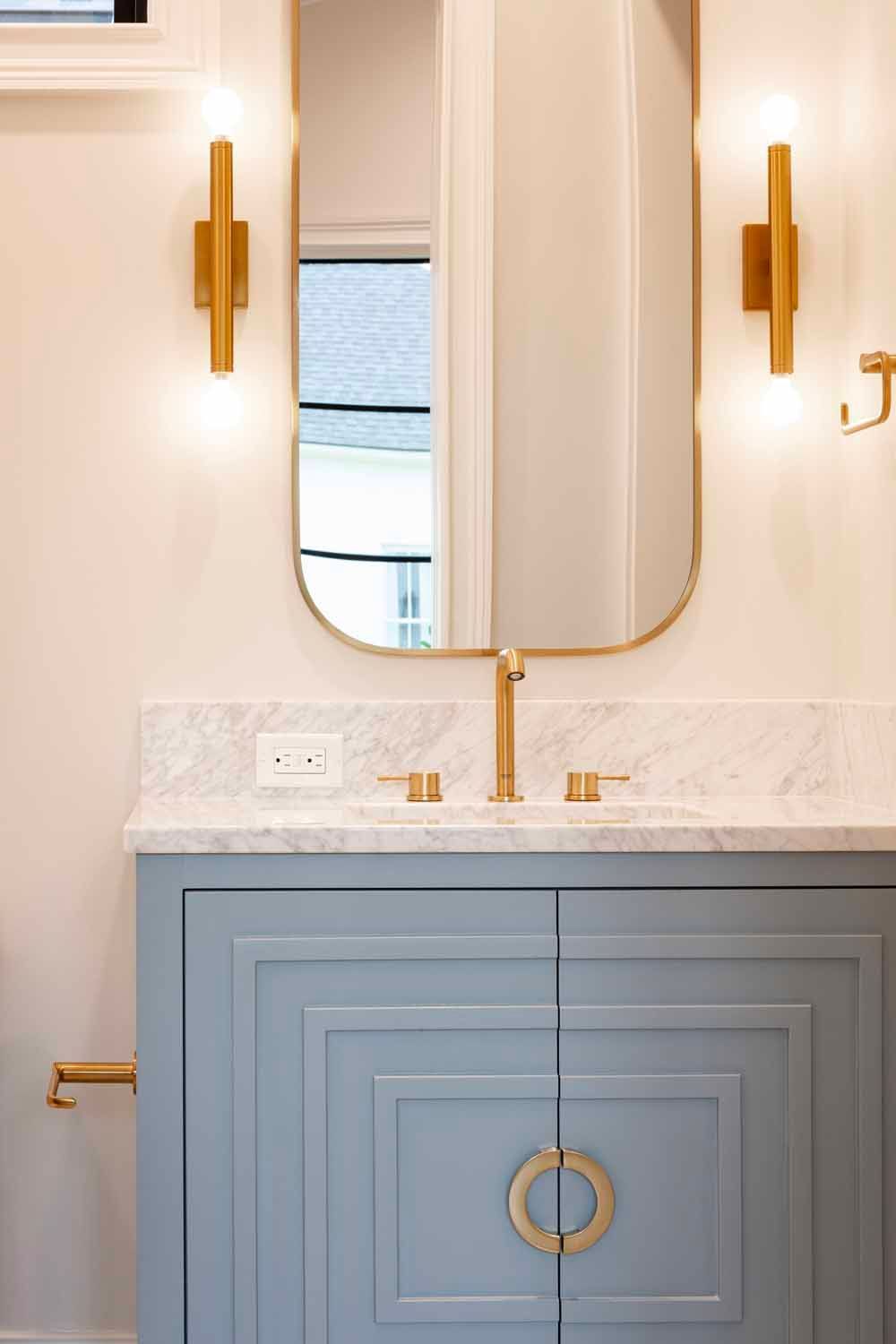 A vanity with a grey cabinet, marble countertop, gold faucet, wall-mounted light fixtures, and an arched gold mirror.