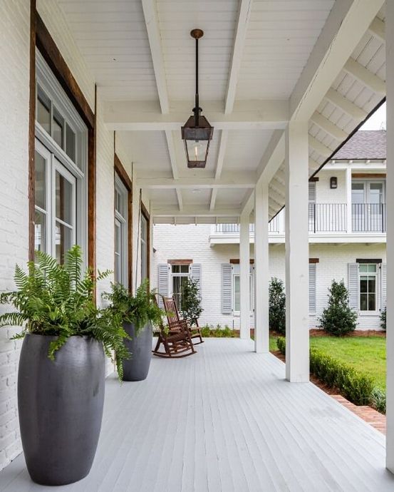 A covered porch with light-painted wood, white pillars, two gray planters with ferns, and two wooden rocking chairs.